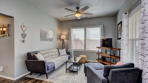 A living room with a sofa, armchairs, a coffee table, a ceiling fan, and a TV stand. - The Red Bench Cottage | Historic Charm • Fenced Yard • Walk to Hyde Park • Central Boise (Boise)