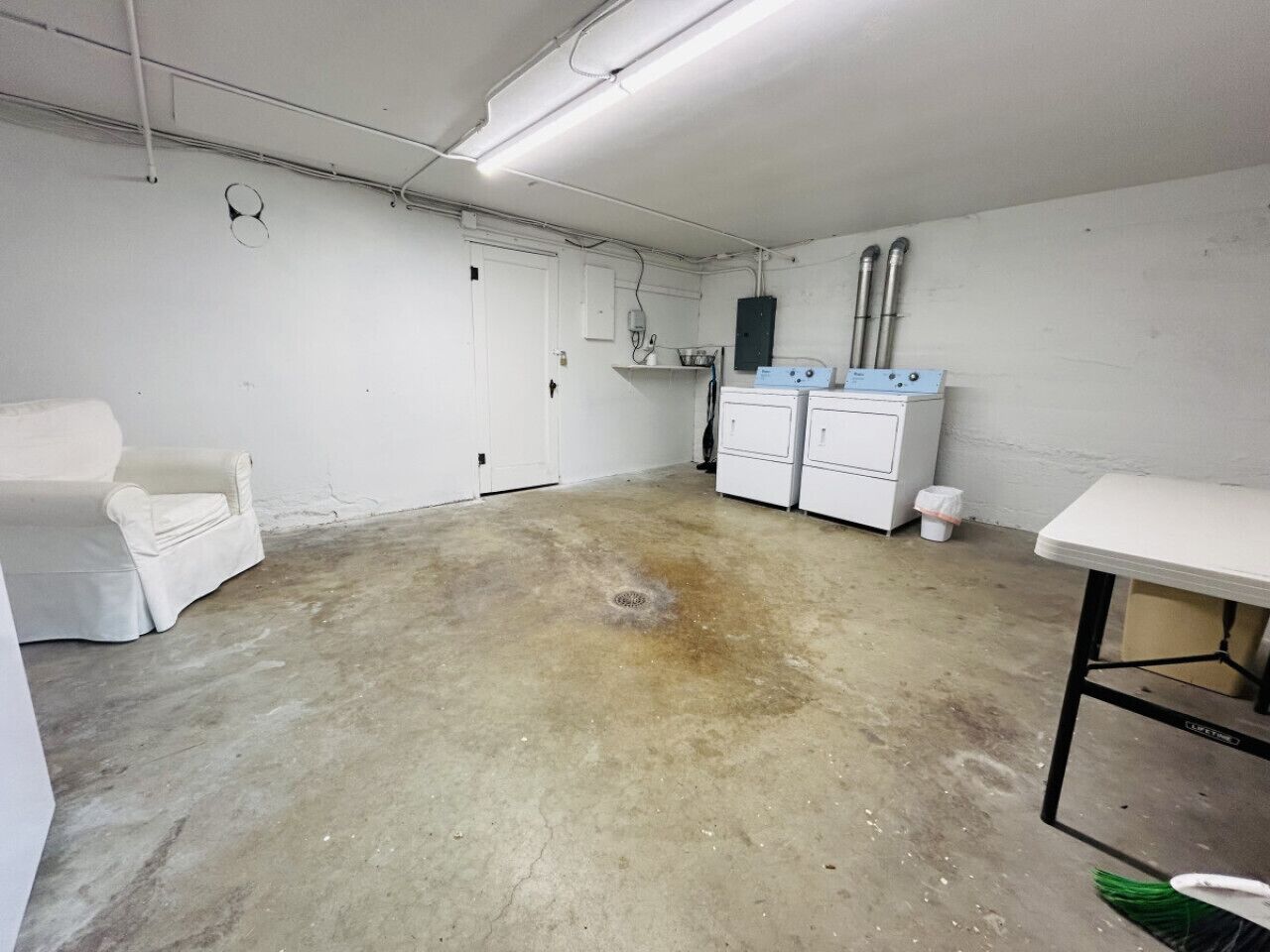 Una habitación amplia con suelo de hormigón, paredes blancas y una mesa blanca con estructura metálica. Se observan dos armarios blancos y una silla blanca.