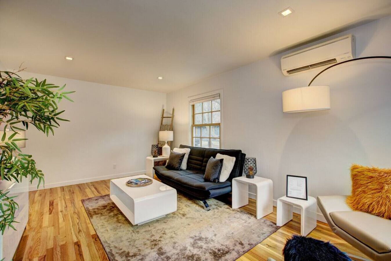 Uma sala de estar moderna com um sofá escuro, mesa de centro branca e uma pequena mesa lateral. Há uma janela grande que oferece luz natural e um aparelho de ar condicionado fixado na parede.