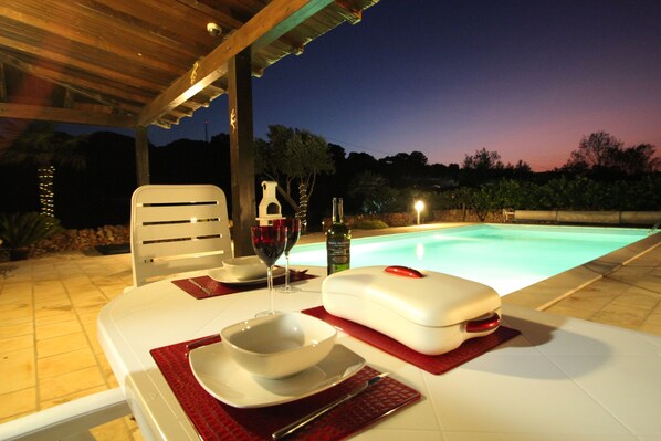 Uma área de refeições ao ar livre com piscina, mesa branca, jogos americanos vermelhos, uma taça de vinho e uma garrafa de vinho.