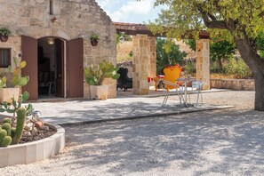 Un patio con un edificio de piedra, plantas de cactus, una silla de metal y una mesa con un objeto rojo encima.