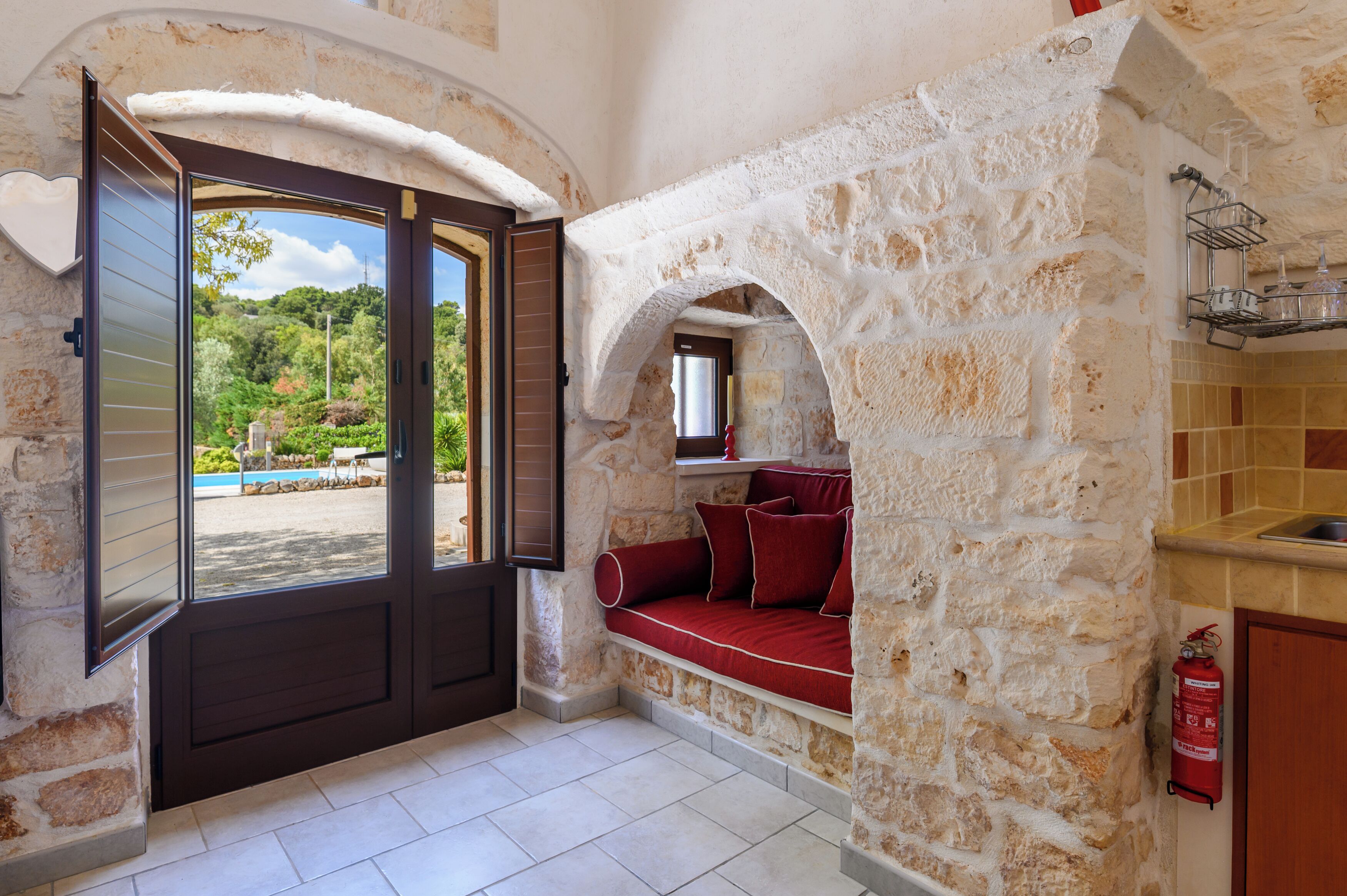 Habitación de paredes de piedra con un banco acolchado rojo, una puerta de madera y vistas a una piscina y follaje exterior.