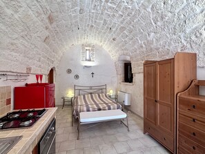 Quarto com cama, mesa de cabeceira, cômoda e cozinha equipada com fogão e armários.