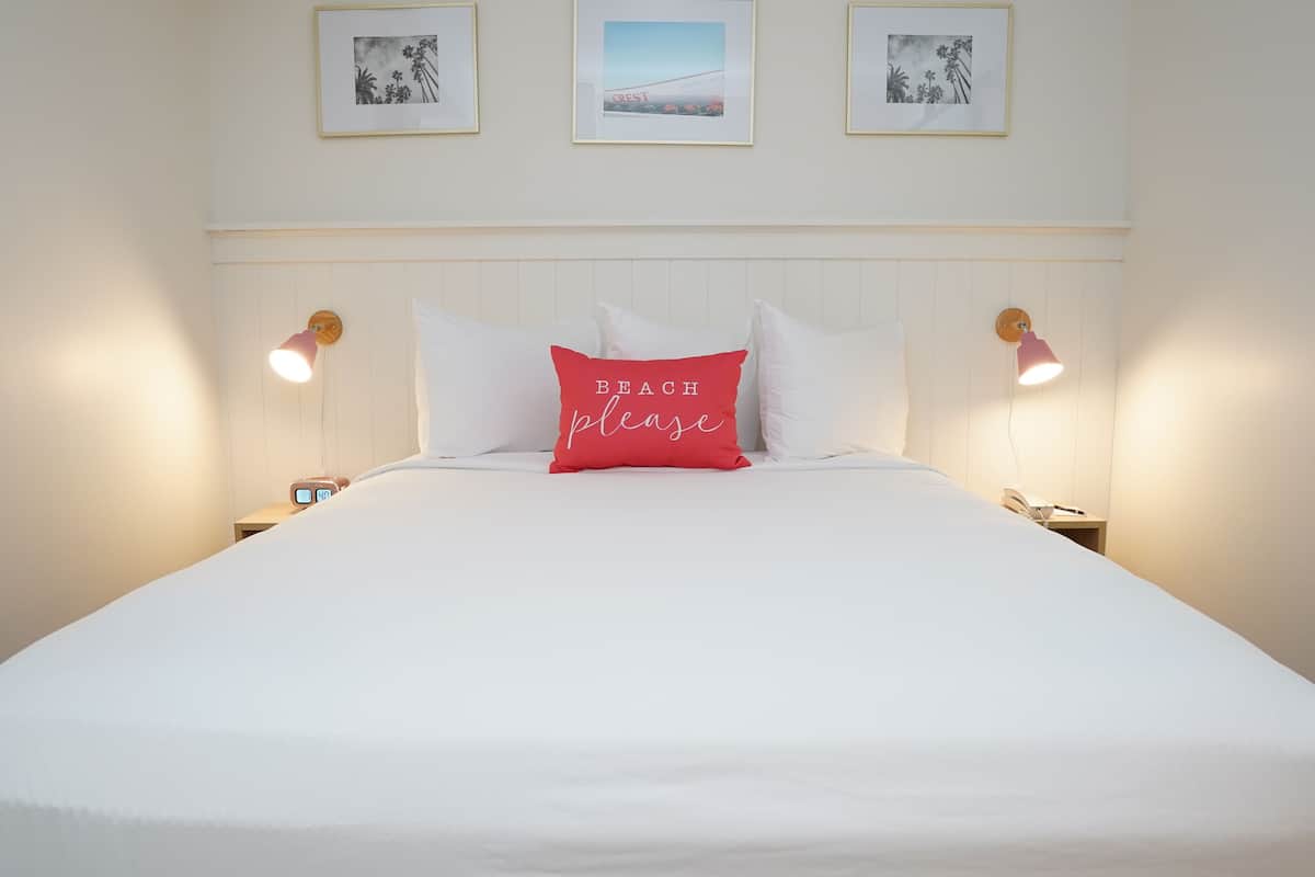 Ein ordentlich bezogenes Bett mit weißer Bettwäsche und einem roten Kopfkissen mit dem Aufdruck "BEACH please". Zwei Nachttischlampen, ein gerahmtes Bild über dem Bett und ein kleiner Nachttisch mit einem Telefon sind ebenfalls zu sehen.