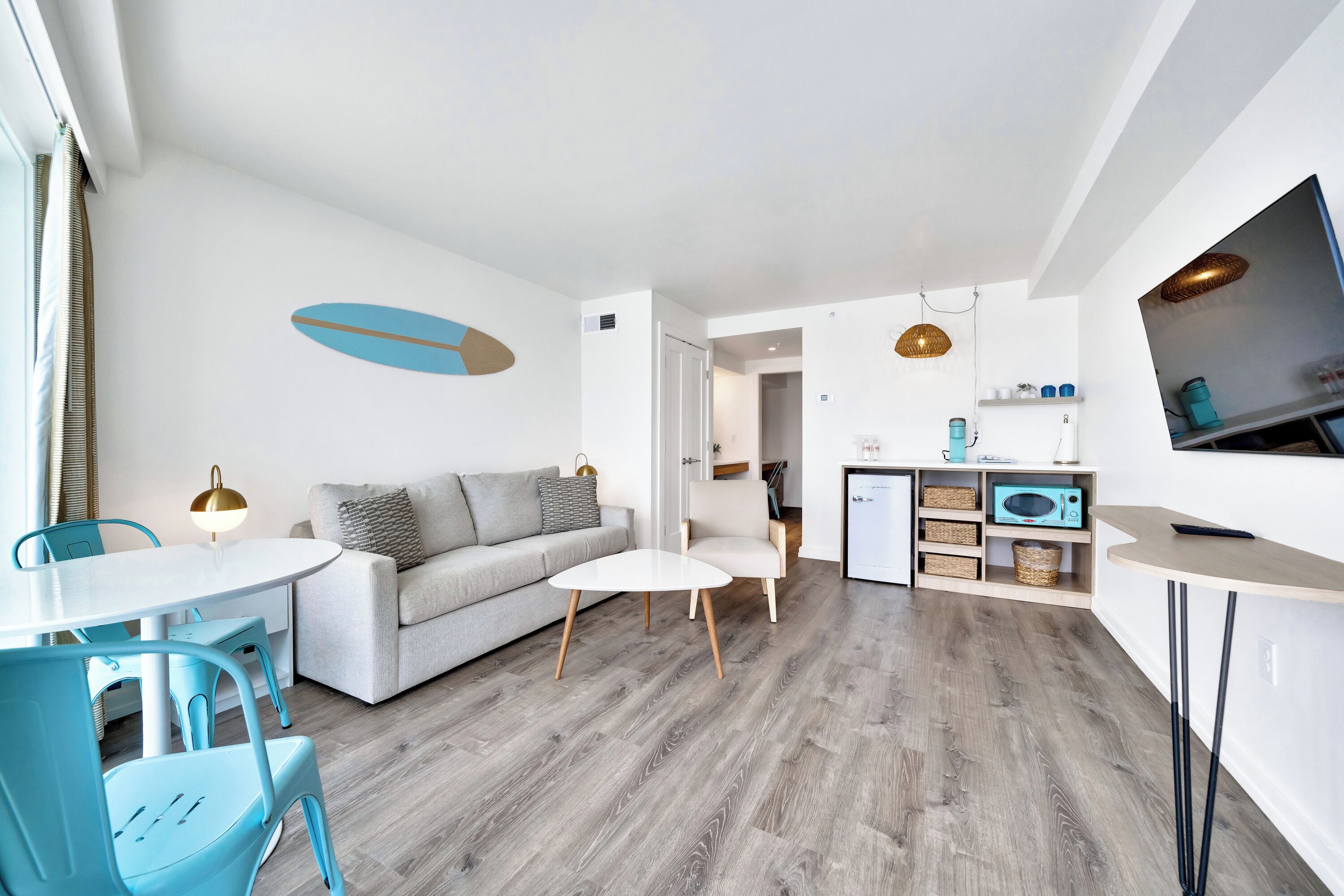 A modern living room with a sofa, a dining table, and chairs. There is a kitchen area with a microwave and shelves.