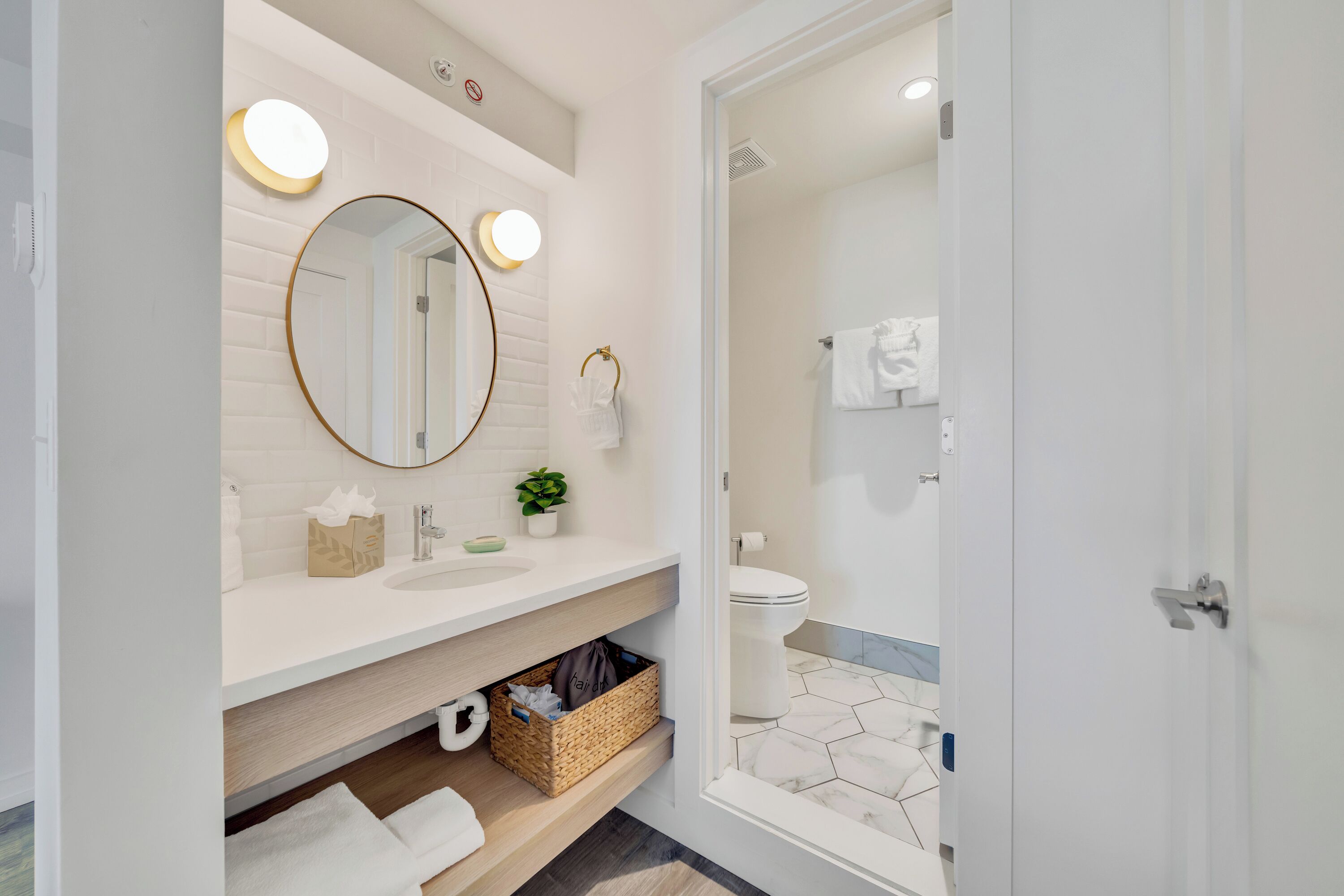 A modern bathroom with a white vanity, a round mirror with lights, a toilet, and a shower area.