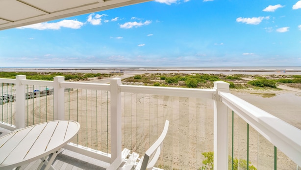 Un balcon avec une table et des chaises, donnant sur une plage, avec un ciel dégagé et une côte lointaine.