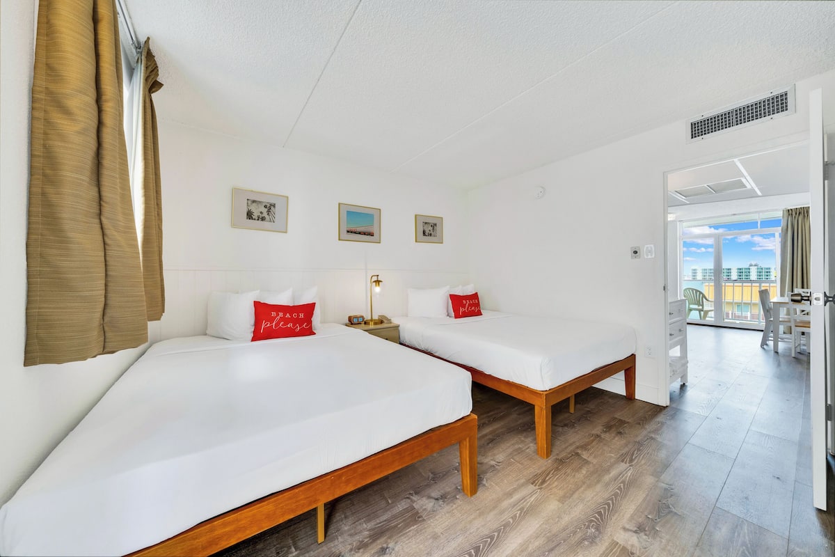 Ein Hotelzimmer mit zwei Einzelbetten, Holz-Kopfteilen, weißer Bettwäsche und roten Kissen. An der Wand hängen Bilder in Rahmen, ein Nachttisch mit einer Lampe und Blick auf die Stadt durch das Fenster.