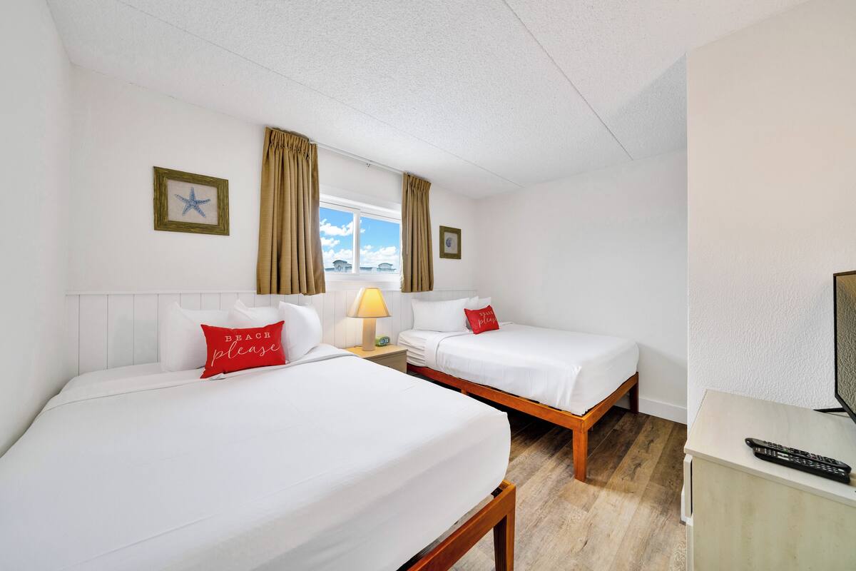 Ein Hotelzimmer mit zwei Betten, einem Nachttisch mit Lampe, einem Fernseher und einem Fenster mit Vorhängen.