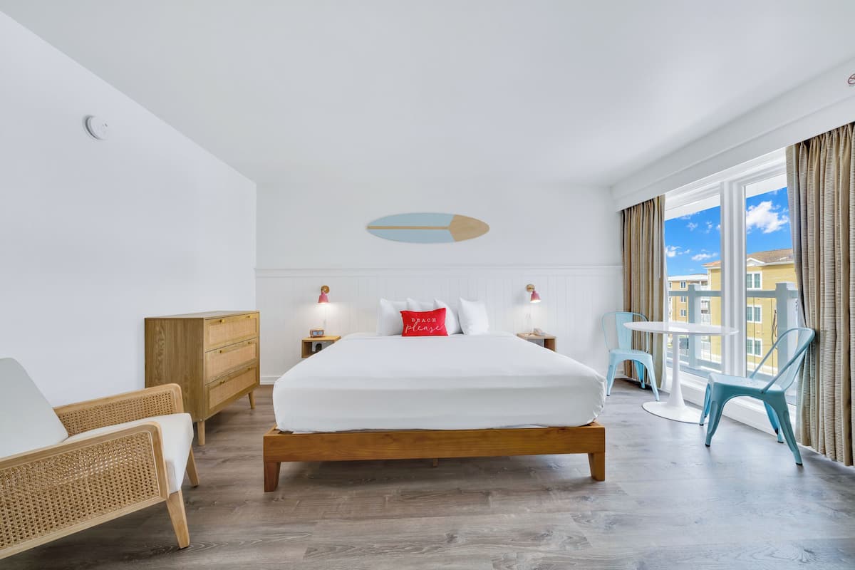 Ein modernes Hotelzimmer mit einem großen Bett, einem Holzkopfstück, zwei Nachttischen mit Lampen, einer kleinen Holzkommode, einem Korbstuhl, einem runden Tisch mit Stühlen und großen Fenstern mit Vorhängen.