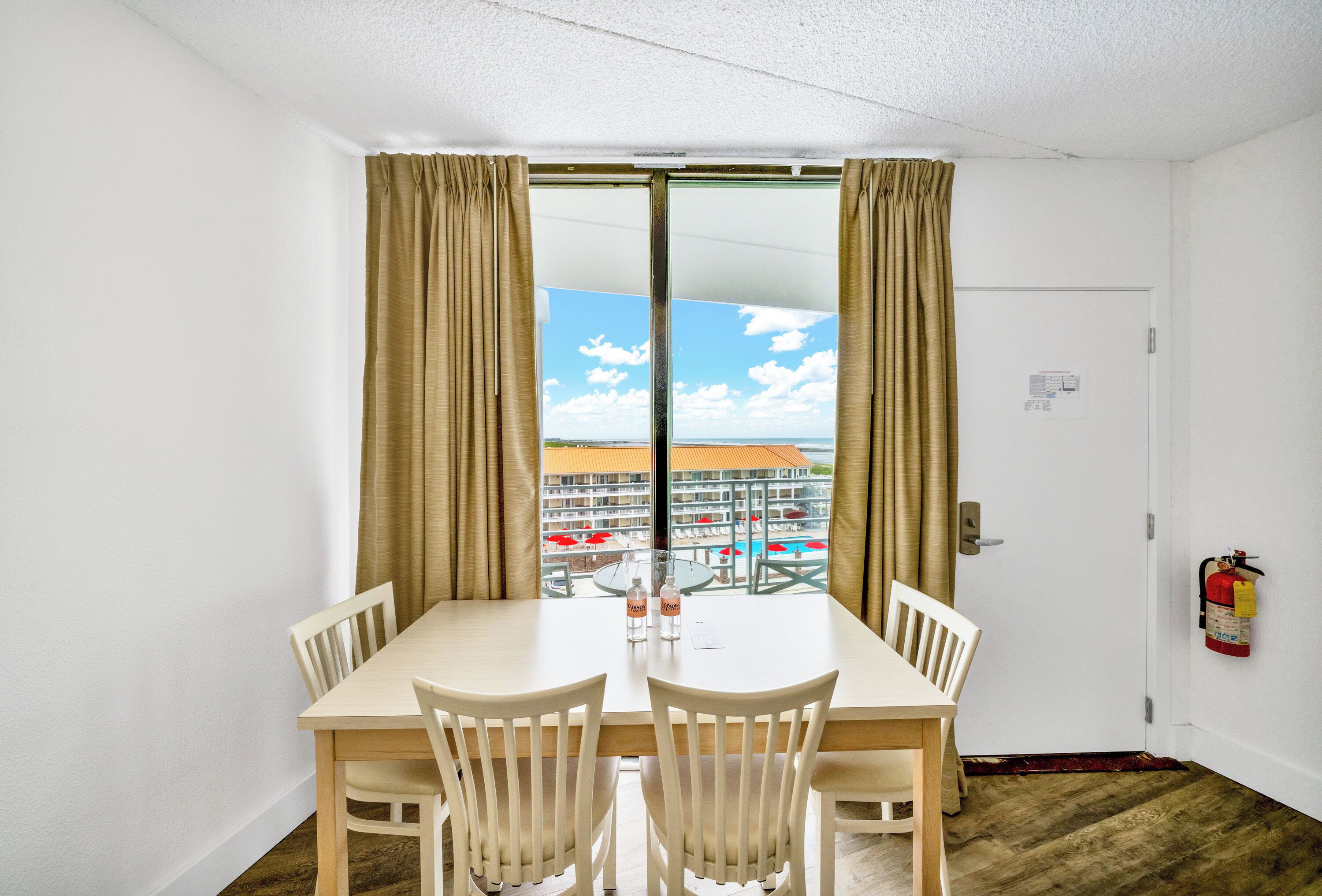 A room with a dining table and chairs, a window with curtains, and a fire extinguisher on the wall.