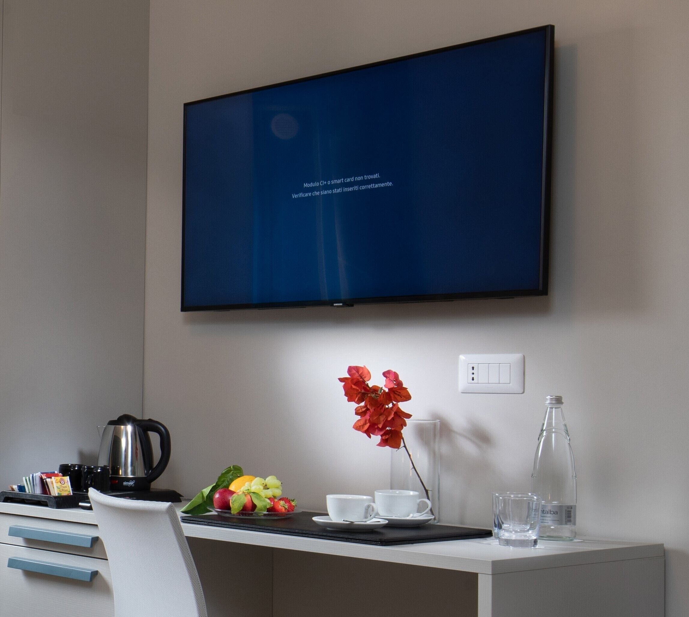 Una habitación de hotel moderna con una televisión de pantalla plana montada en la pared, un escritorio con cafetera, fruta, tazas y un florero con flores.