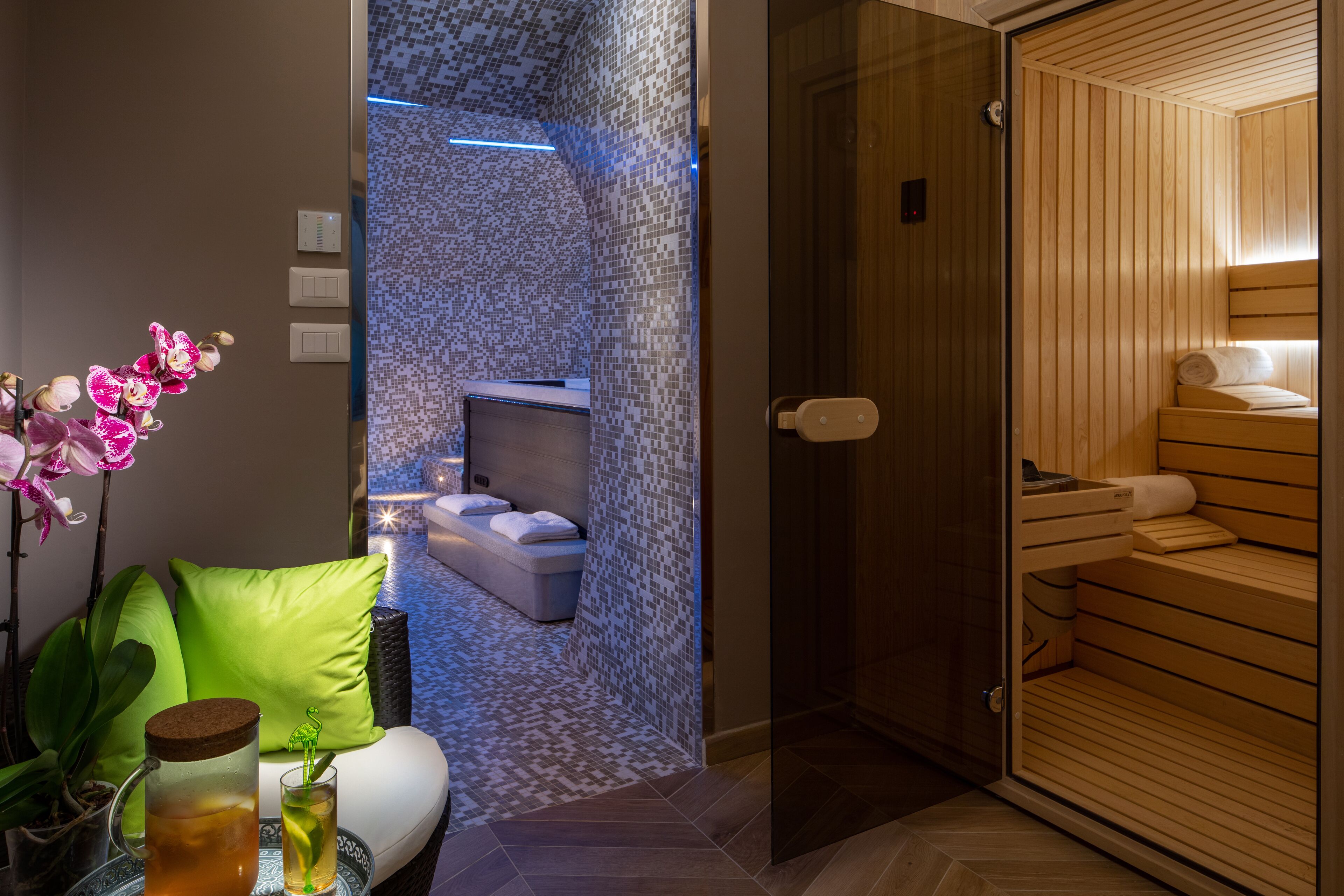 A modern bathroom with a glass shower enclosure, a wooden sauna, and a seating area with a green cushion.