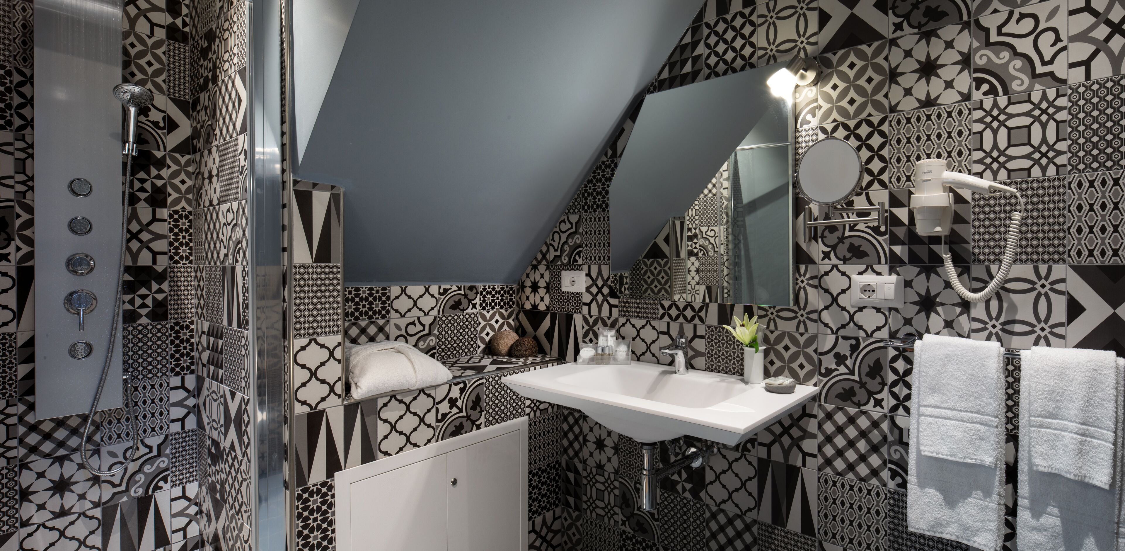 Une salle de bain moderne avec un carrelage aux motifs géométriques, un lavabo blanc, une douchette à main, un porte-serviettes avec des serviettes et un miroir avec un éclairage intégré.