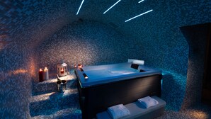 A modern spa room with a large bathtub, a small seating area, and a tiled wall. - Casa Blandano (Castelmola)