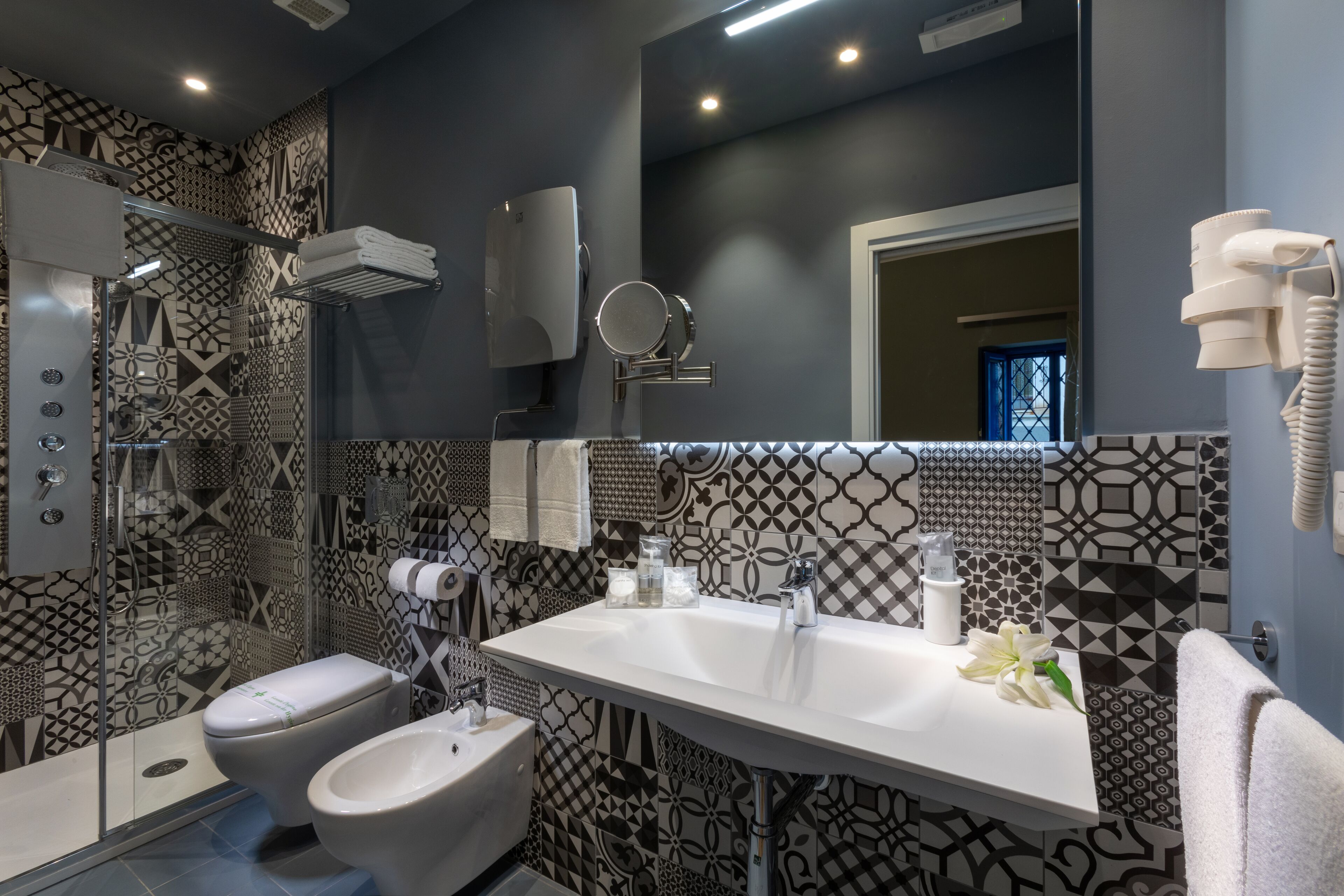 Un baño moderno con un lavamanos blanco, dos inodoros, ducha y azulejos estampados.