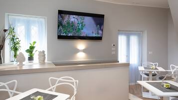A modern dining area with white chairs and tables, a flat-screen TV mounted on the wall, and a window with sheer curtains.