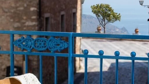 A wooden table with a white cup, a book, and an apple on a balcony with a blue railing. - Casa Blandano (Castelmola)