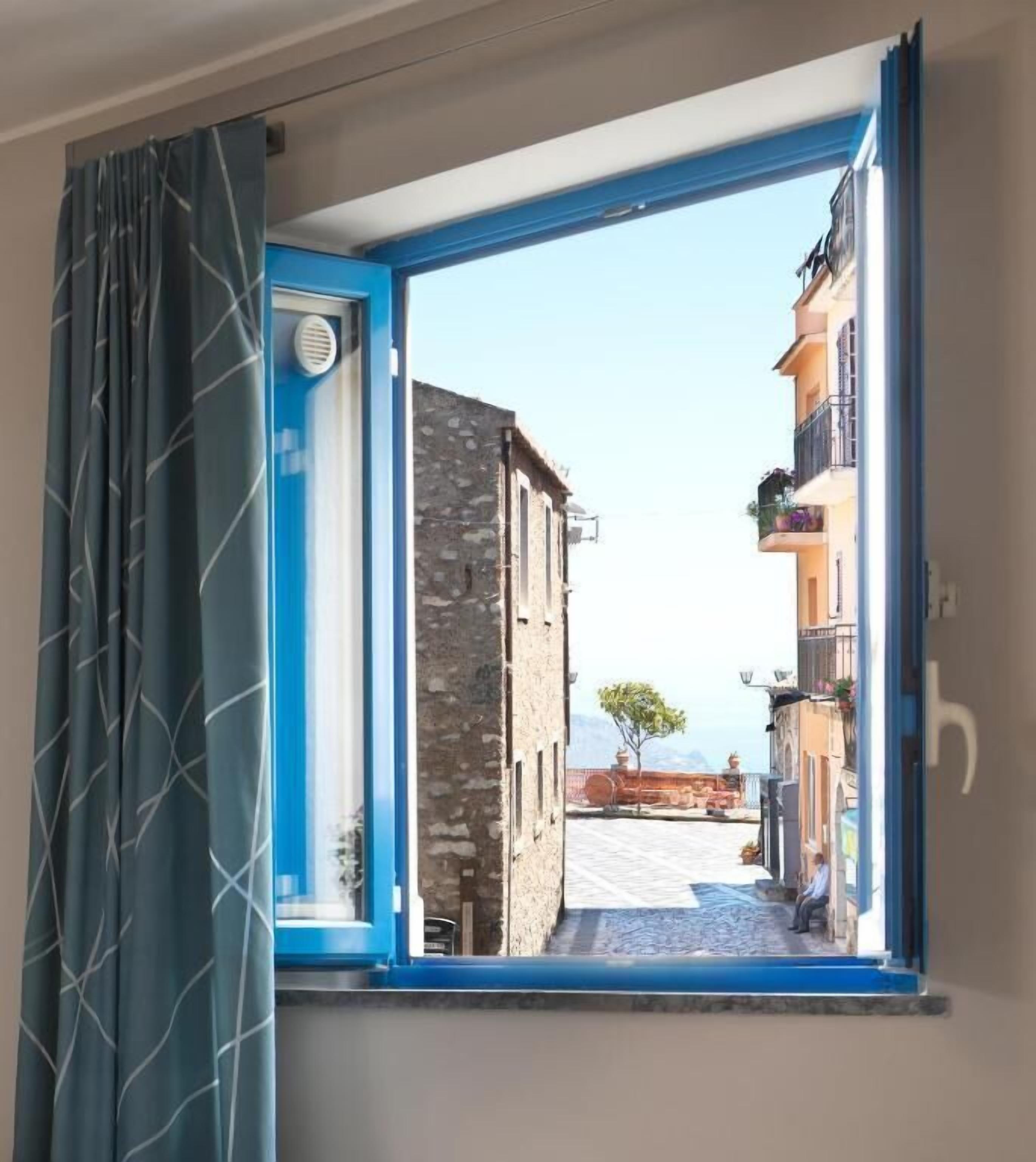 A window with a view of a narrow street with buildings, a tree, and a clear sky.