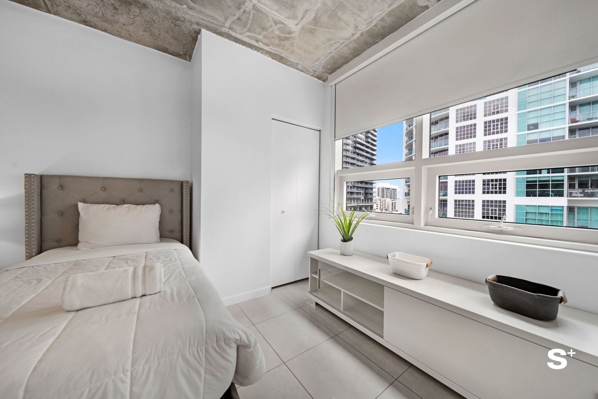 Une chambre moderne avec un grand lit, un banc intégré, une fenêtre donnant sur la ville et un plafond en béton.