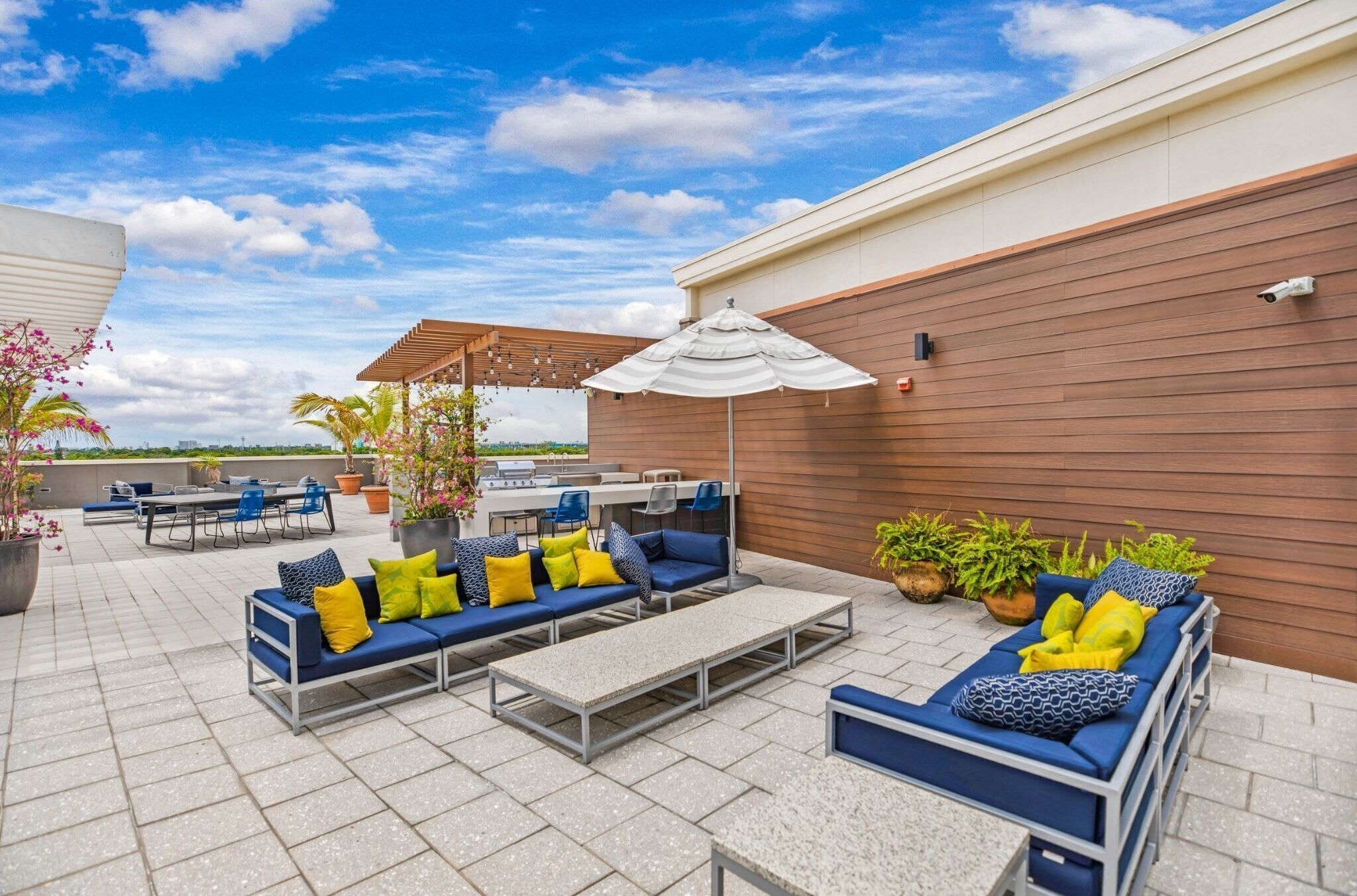 Une terrasse extérieure avec des sièges rembourrés aux couleurs bleu et jaune, une table et des plantes en pot.