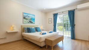 A bedroom with a bed, a wicker bench, a painting, a window with curtains, and an air conditioning unit.