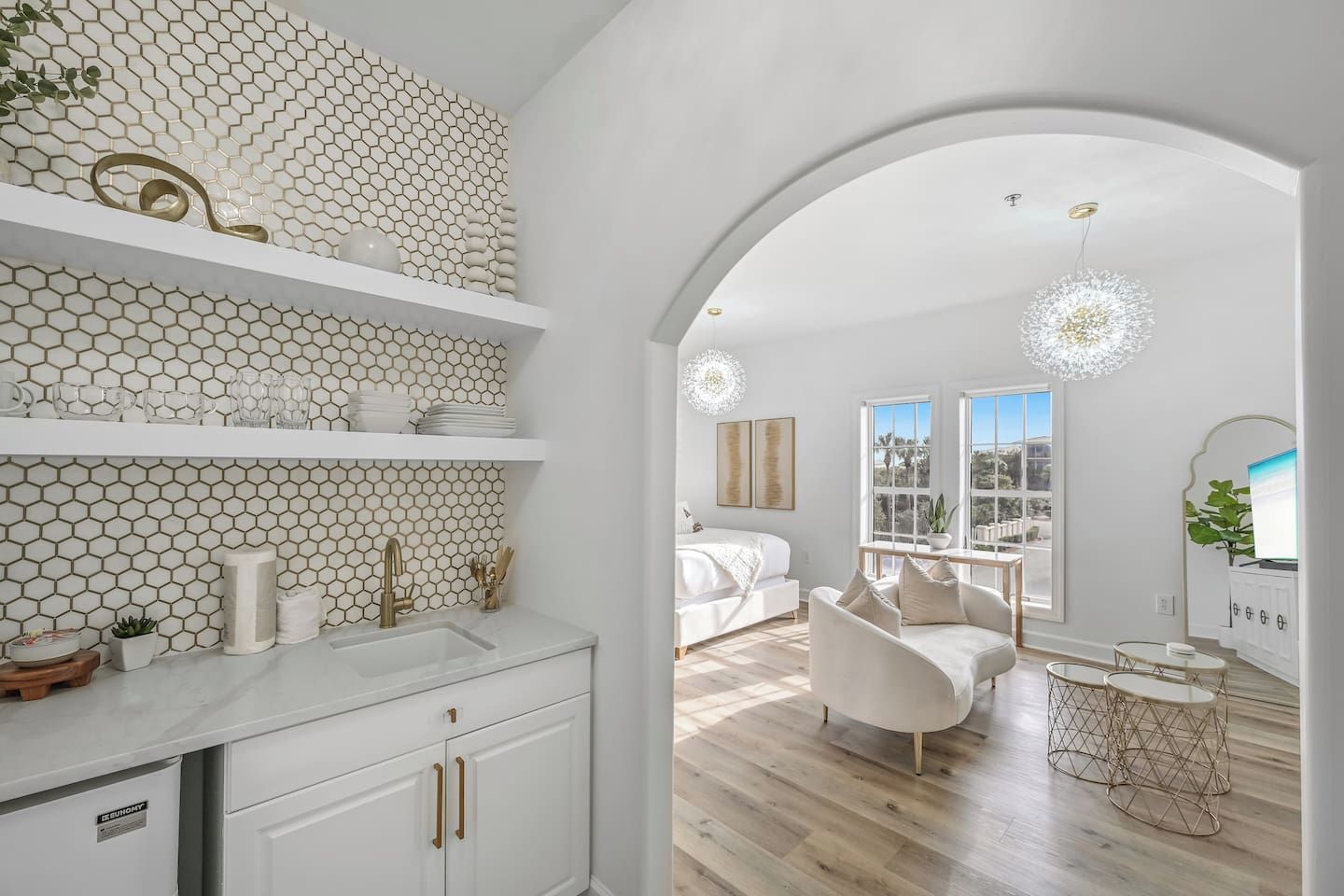 A modern interior with a kitchen area, white cabinetry, a dining area with a sofa and table, and a large arched doorway leading to a bedroom with a view.