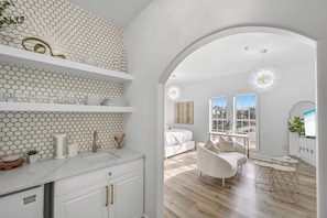 A modern interior with a kitchen area, white cabinetry, a dining area with a sofa and table, and a large arched doorway leading to a bedroom with a view.