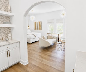 A modern interior with a white sofa, a dining table with chairs, and a kitchen area with white cabinets and a hexagonal tile backsplash.