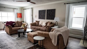 Ein Wohnzimmer mit einer braunen Sofagarnitur, einem Couchtisch und einer Stehlampe. An den Wänden hängen zwei Kunstwerke und es gibt einen Deckenventilator.