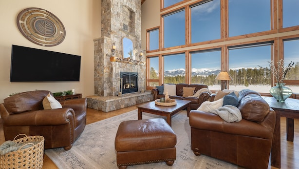 A spacious living room with leather sofas, a stone fireplace, large windows, and a flat-screen TV. - Family-friendly cabin near Winter Park w/ hot tub (Fraser)