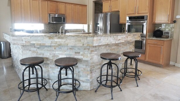 A kitchen with a stone island, wooden cabinets, stainless steel appliances, and four stools. - Rancho Mirage Home With Large Pool (Rancho Mirage)