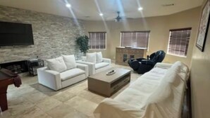 A spacious living room with a sofa, a coffee table, and a TV mounted on a stone wall. - Rancho Mirage Home With Large Pool (Rancho Mirage)