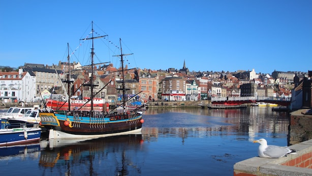 Ein Hafen mit einem großen Segelschiff, das anlegt, umgeben von Gebäuden und ruhigem Wasser.