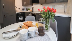 Eine moderne Küche mit dunklen Schränken, einer Mikrowelle und einem Spülbecken. Auf dem Tisch befinden sich Croissants, Kaffeetassen und ein Kaffeekrug.
