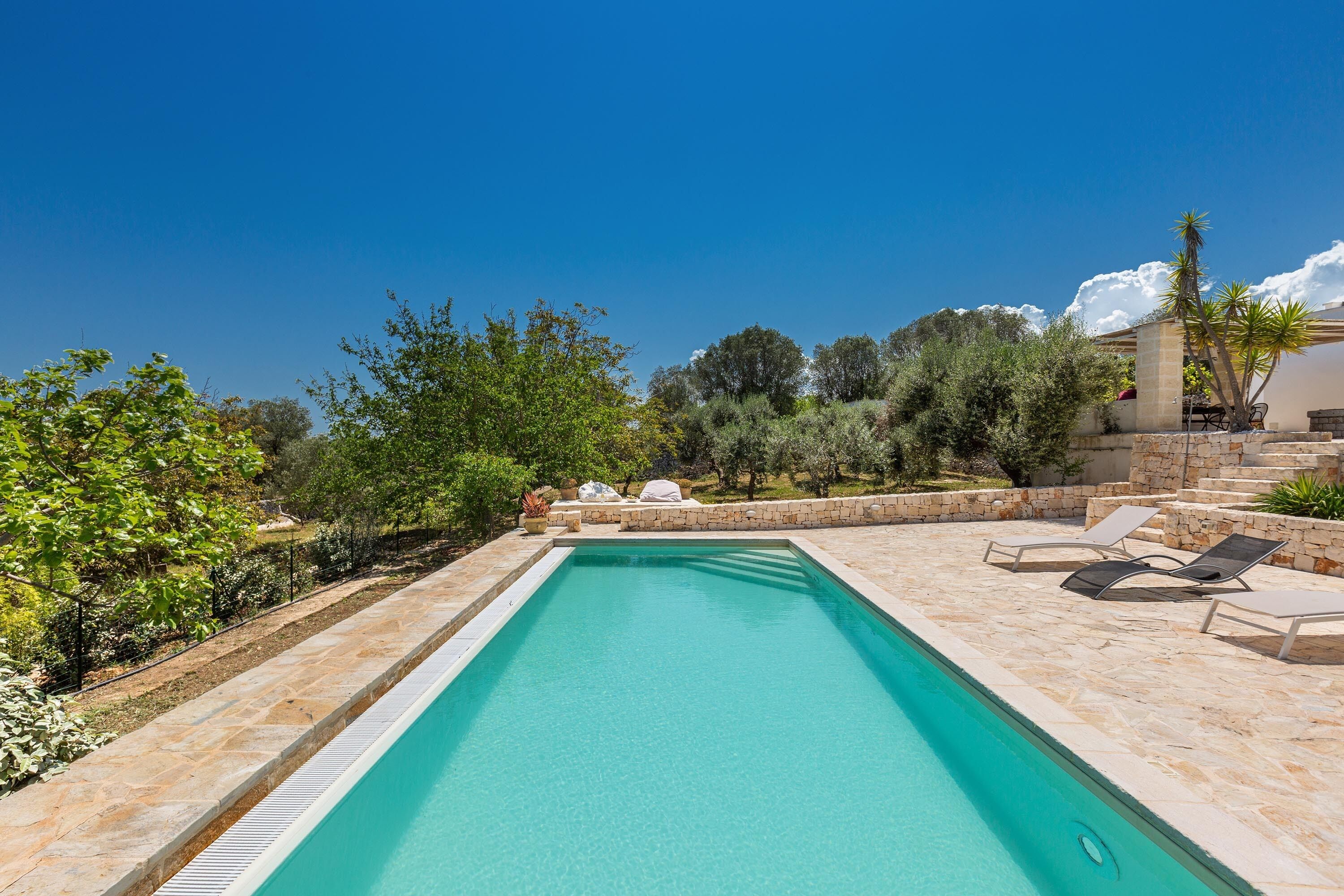 A rectangular swimming pool with a stone border, surrounded by a stone patio with lounge chairs and a covered seating area.