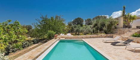 A rectangular swimming pool with a stone border, surrounded by a stone patio with lounge chairs and a covered seating area.