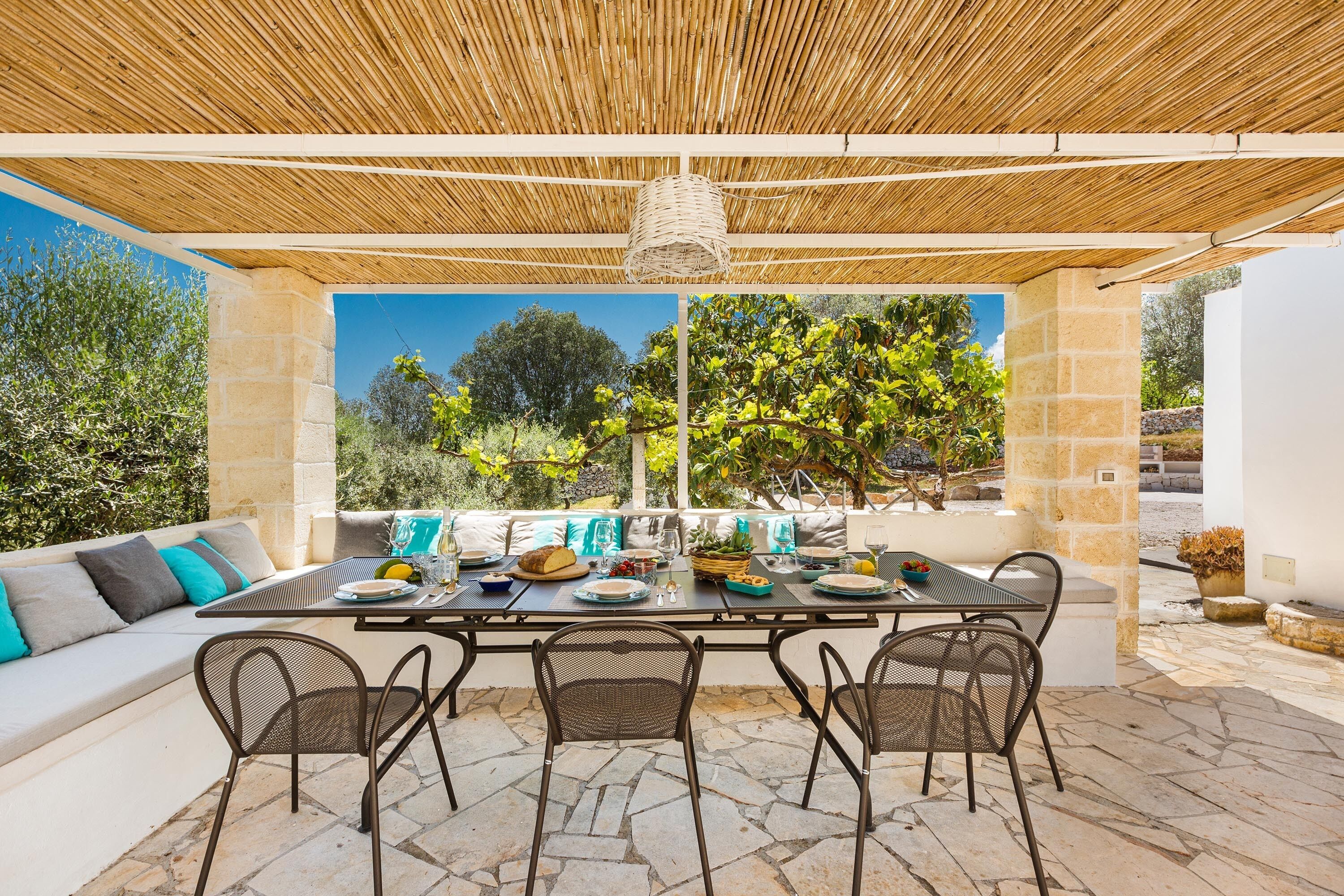 An outdoor dining area with a table set for four, surrounded by wicker chairs, under a bamboo roof.