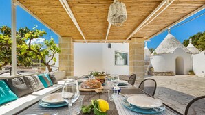 An outdoor dining area with a table set for two, a wicker basket hanging from the ceiling, and a view of a white building with a conical roof.