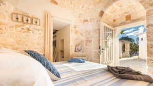 A bedroom with stone walls, a bed with striped bedding, a fan, and a view of the outside.