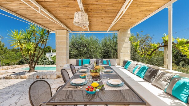 An outdoor dining area with a long table set for a meal, surrounded by cushioned seating, and a view of a pool and greenery.