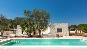 A pool area with a stone building, outdoor seating, and trees. - 2499 Trulli La Meringa by Perle di Puglia (Ostuni)