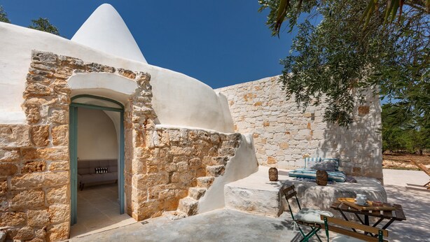 A stone building with a white exterior, a doorway, and a seating area with a table and chairs. - 2499 Trulli La Meringa by Perle di Puglia (Ostuni)