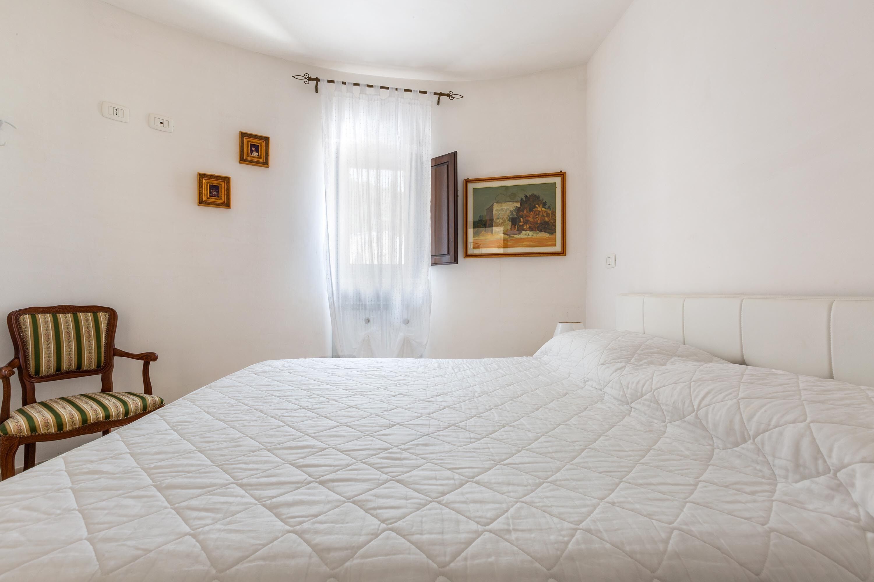 Un lit bien fait, recouvert d’une couette blanche matelassée, une chaise en bois avec un coussin à rayures et un tableau accroché au mur.