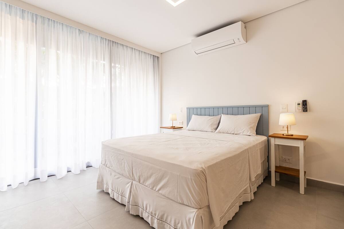 A neatly made bed with a blue headboard, two bedside tables with lamps, and a large window with sheer curtains.