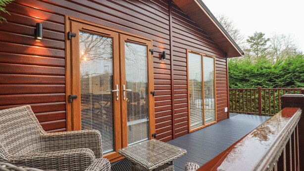 A wooden cabin with a covered deck, wicker furniture, and a glass door. - 25 Thirlmere (Windermere)