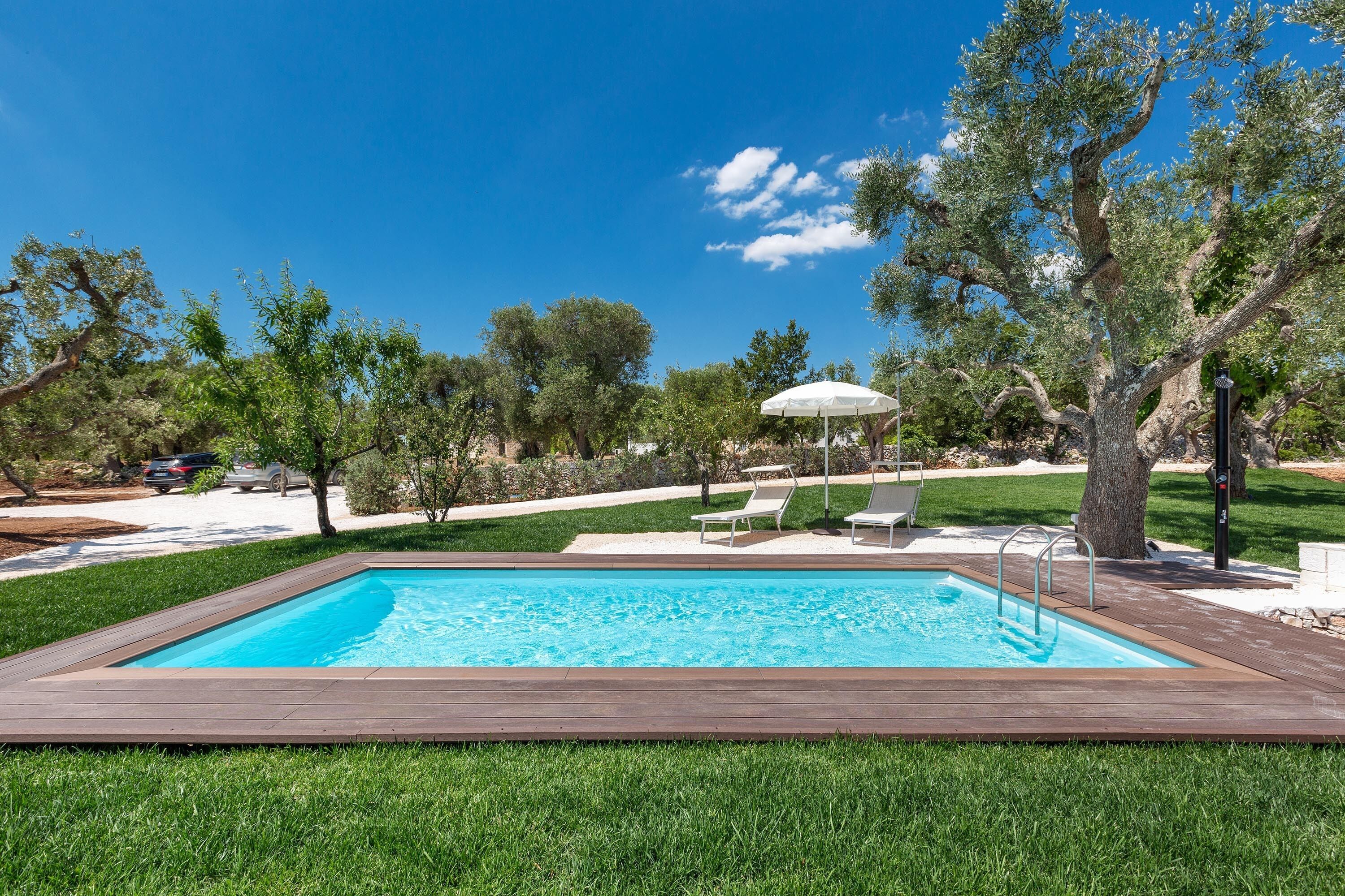 A rectangular swimming pool with a wooden deck, surrounded by green grass and trees.