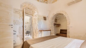 Een slaapkamer met stenen muren, een bed met een beige en witte dekbedovertrek, een houten nachtkastje en een klein houten plankje.