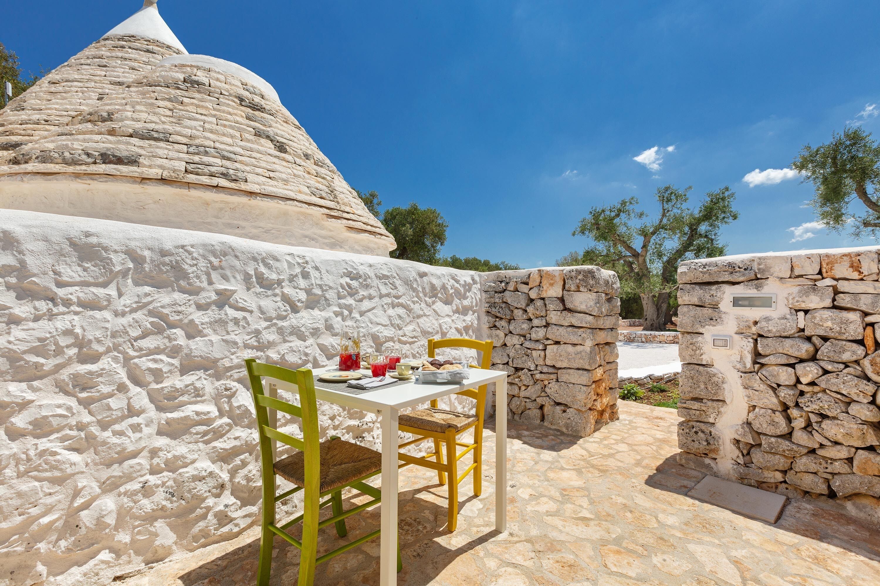 Un espacio al aire libre con paredes de piedra, con mesas y sillas, una estructura abovedada de piedra y un cielo azul despejado.