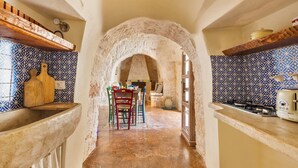 A narrow kitchen with stone walls, a tiled backsplash, and a central archway leading to a dining area with colorful chairs. - 2530 Trullolive by Perle di Puglia (Ostuni)